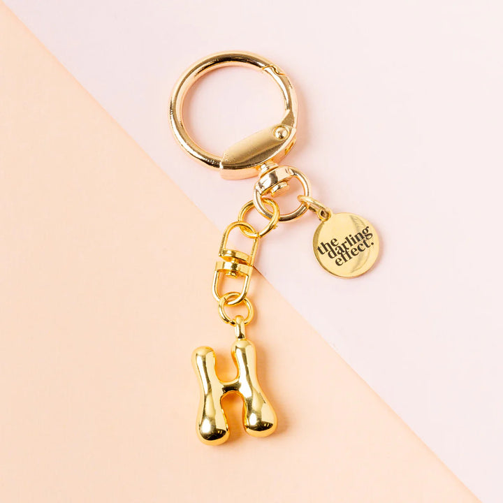 Gold-plated keychain by The Darling Effect featuring a shiny metallic balloon-style letter H charm and a branded circular tag on a diagonal split pink and cream background