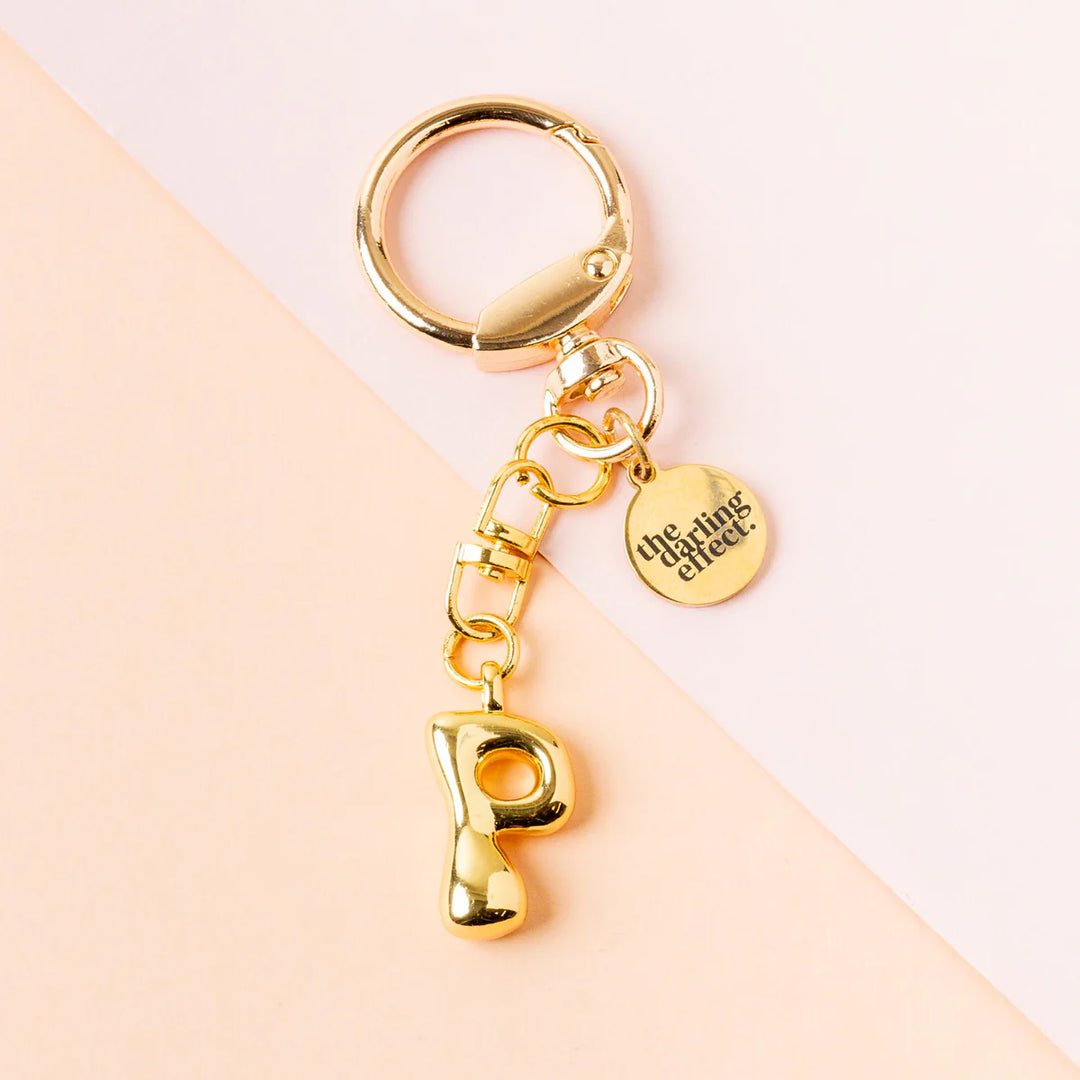 Product photograph by The Darling Effect of a polished gold keychain on a diagonal pink background showcasing a puffy bubble-style letter P charm and a round brand-engraved disk