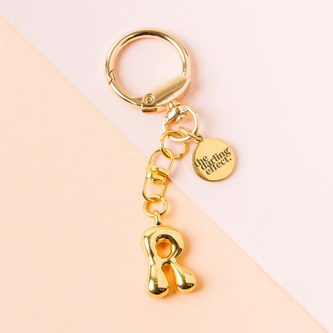 Gold-plated metal keychain by the darling effect featuring a puffy bubble-style letter R charm and an engraved circular logo tag on a split pastel pink and peach background