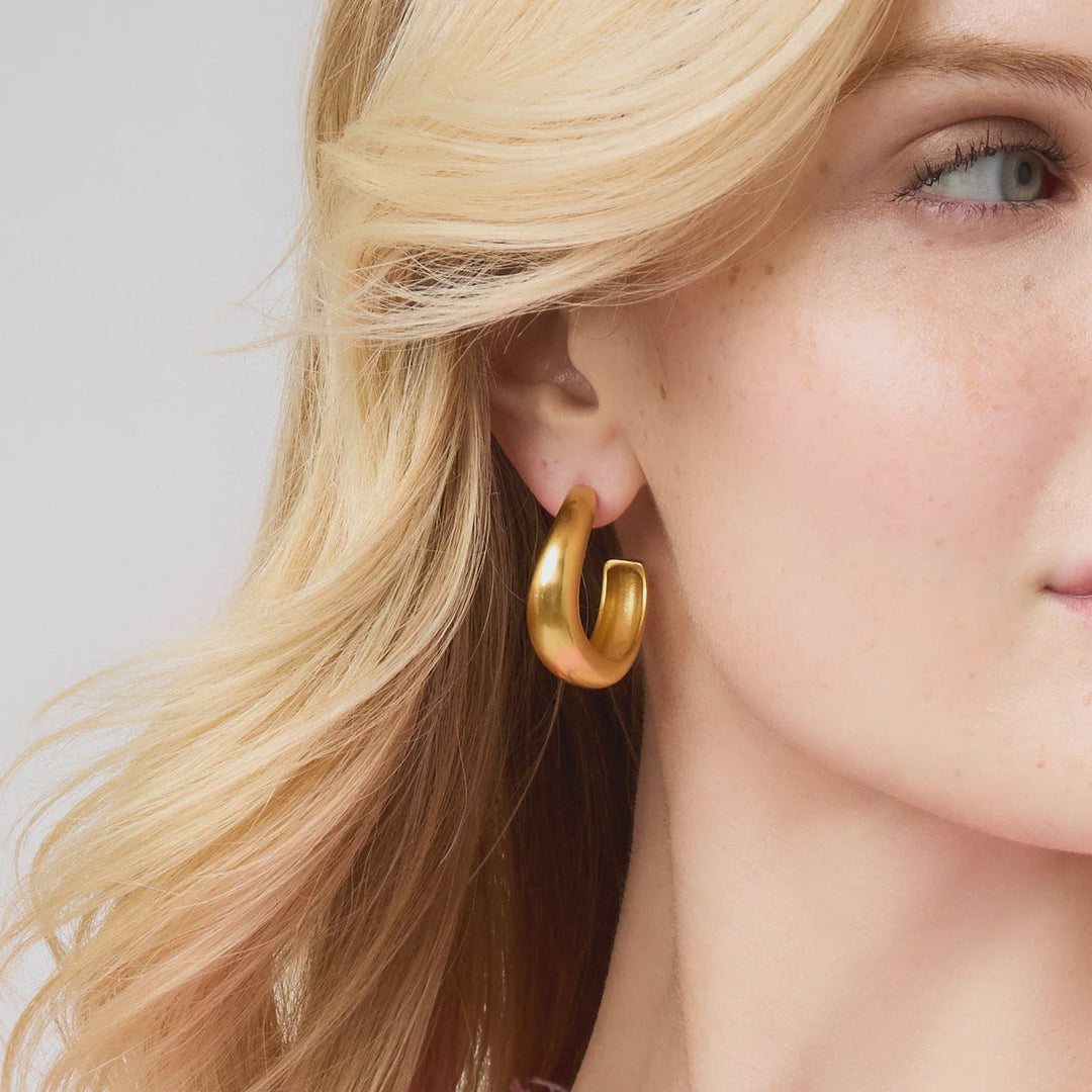 Studio photograph of a blonde woman featuring a thick matte-finished gold hoop earring with a sculpted organic shape and light-catching texture set against her wavy hair and freckled skin