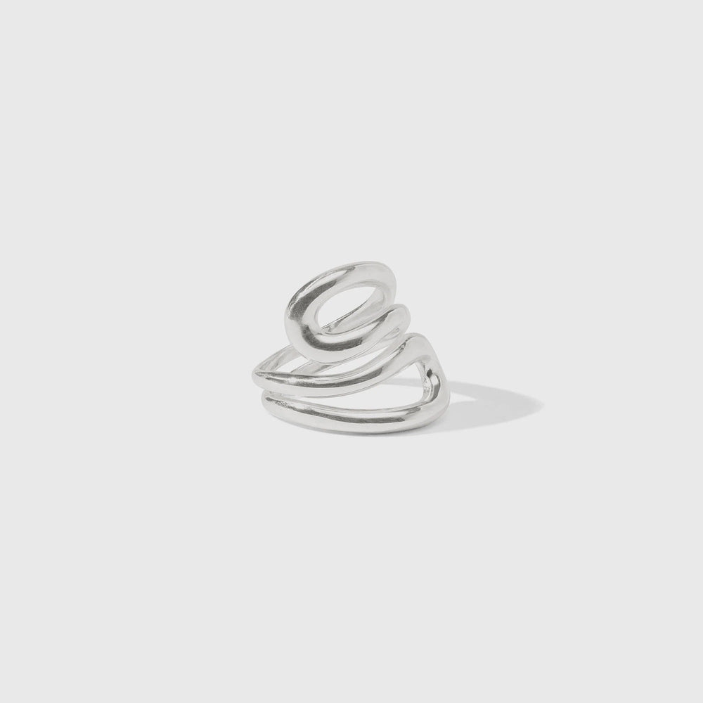 Polished silver sculptural ring by Celine featuring a fluid triple-coil design with overlapping metallic bands and a soft drop shadow against a minimalist gray background