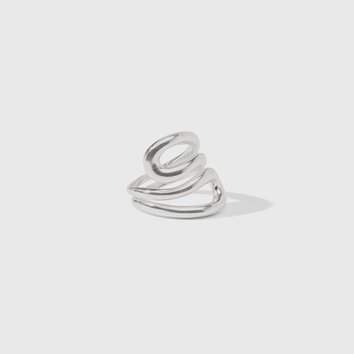 Polished silver sculptural ring by Celine featuring a fluid triple-coil design with overlapping metallic bands and a soft drop shadow against a minimalist gray background