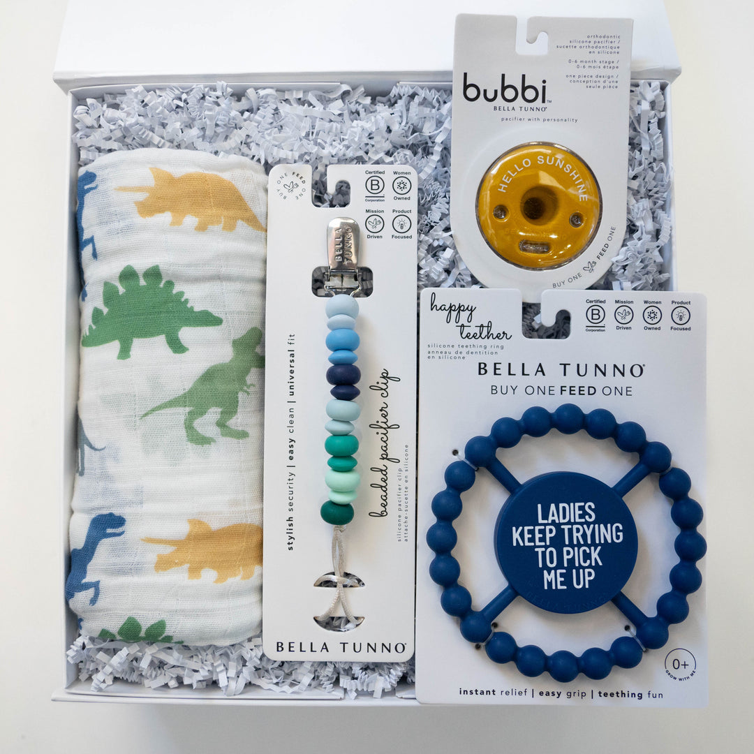 Studio photograph by Bella Tunno of a baby gift set in a white box featuring a colorful dinosaur print muslin swaddle, a multi-tone blue beaded pacifier clip, and a navy blue teething ring with the text Ladies Keep Trying To Pick Me Up