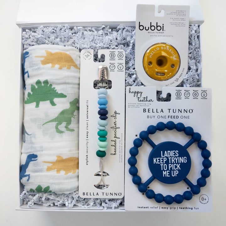 Studio photograph by Bella Tunno of a baby gift set in a white box featuring a colorful dinosaur print muslin swaddle, a multi-tone blue beaded pacifier clip, and a navy blue teething ring with the text Ladies Keep Trying To Pick Me Up