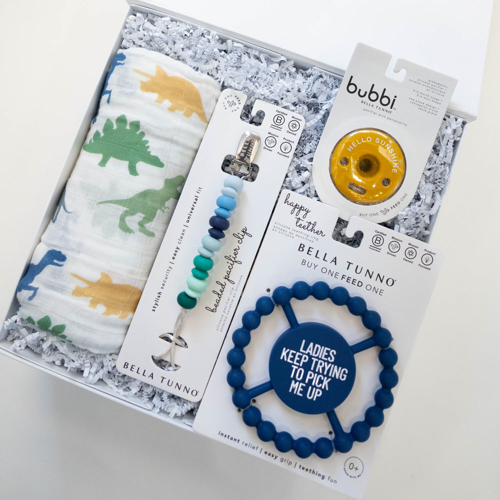 Photograph of a Bella Tunno baby gift set featuring a colorful dinosaur print swaddle, a gradient blue beaded pacifier clip, and a navy teether with the phrase Ladies Keep Trying To Pick Me Up nestled in a white box with shredded paper
