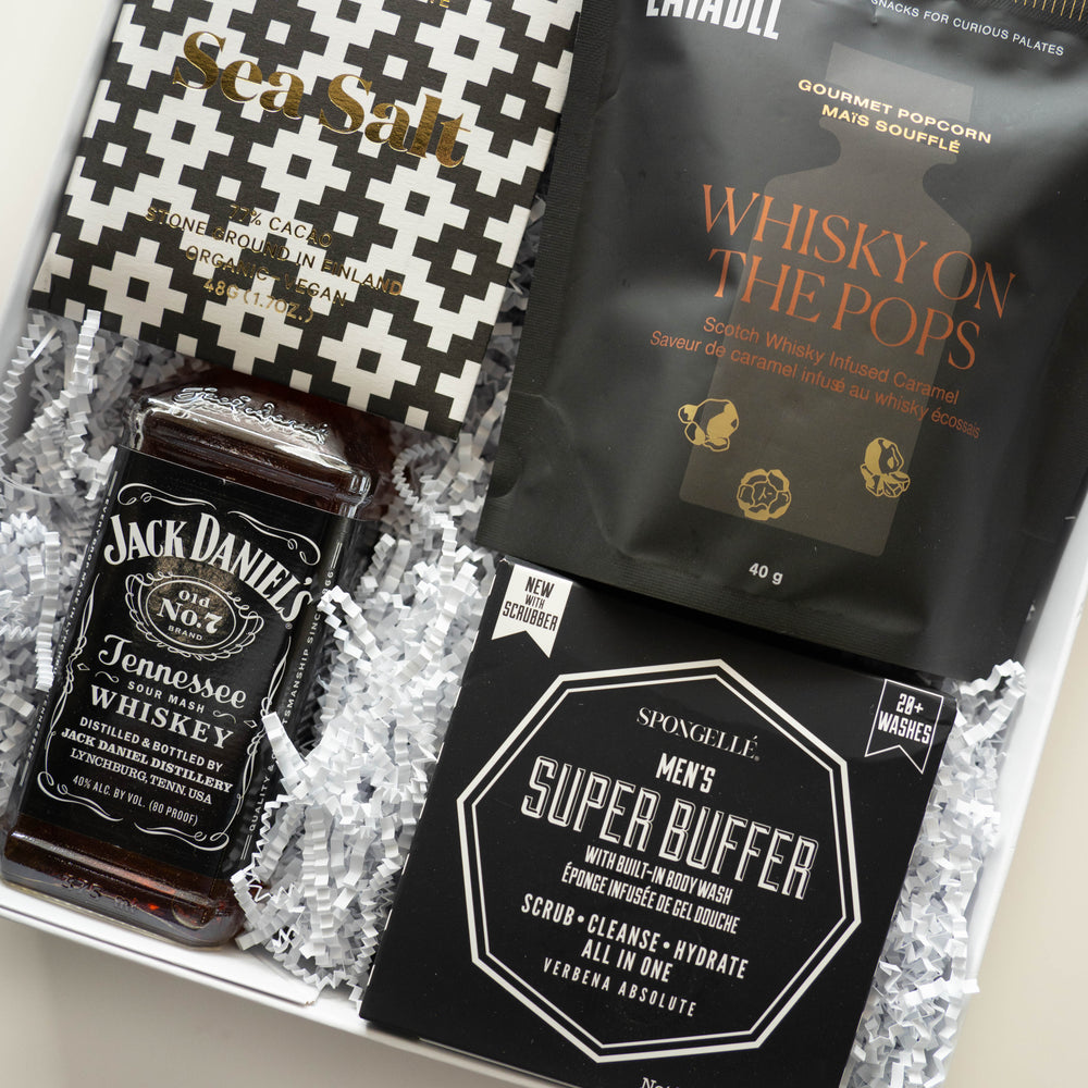 Digital photograph of a men's gift set featuring a Jack Daniel's whiskey bottle and Spongellé Super Buffer accented by a geometric black-and-white chocolate wrapper and copper-toned popcorn packaging on white shredded paper filler