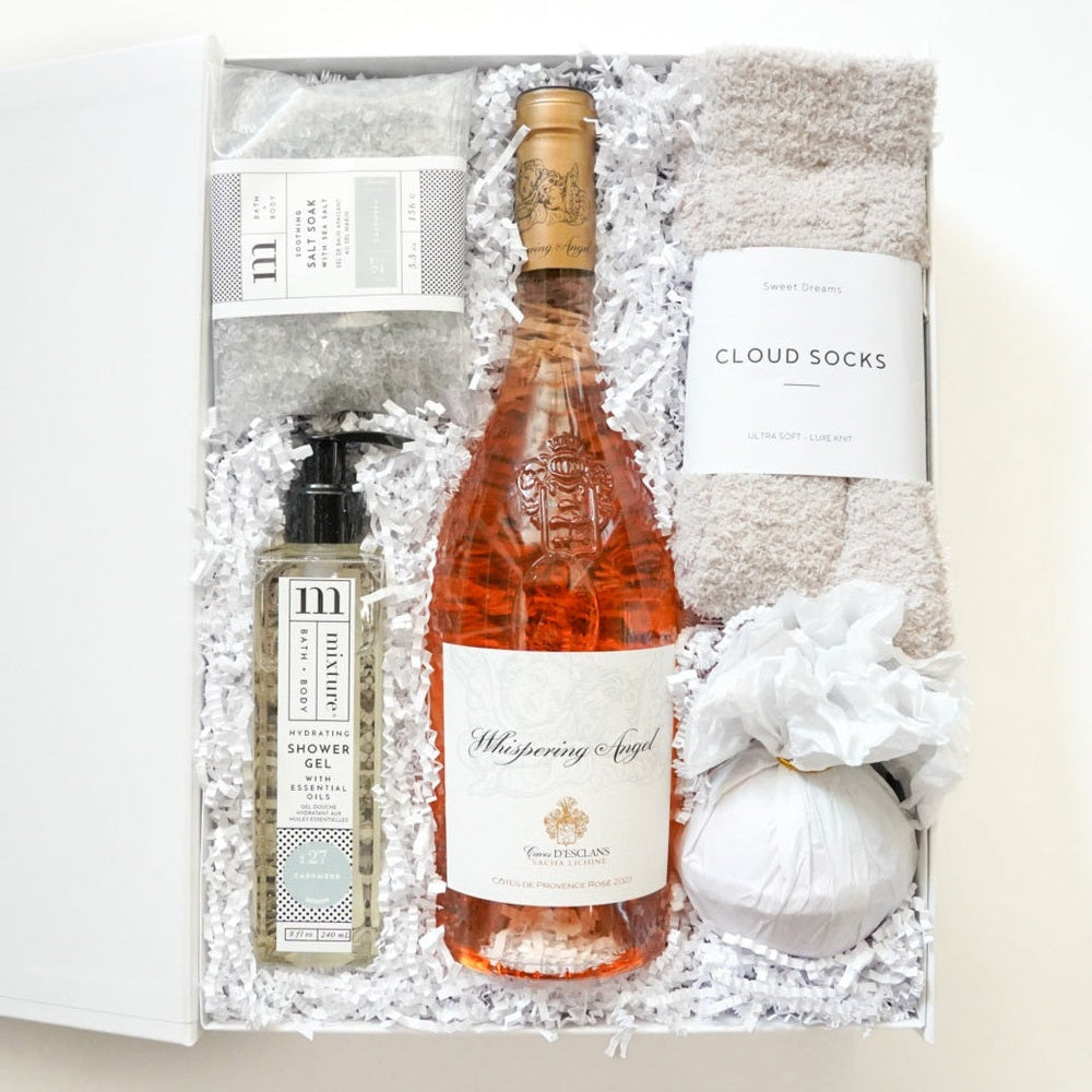 Photograph of a luxury gift box featuring a central bottle of peach-hued Whispering Angel Rosé wine, fuzzy beige Cloud Socks, and Mixture brand bath products resting on white crinkled paper filler