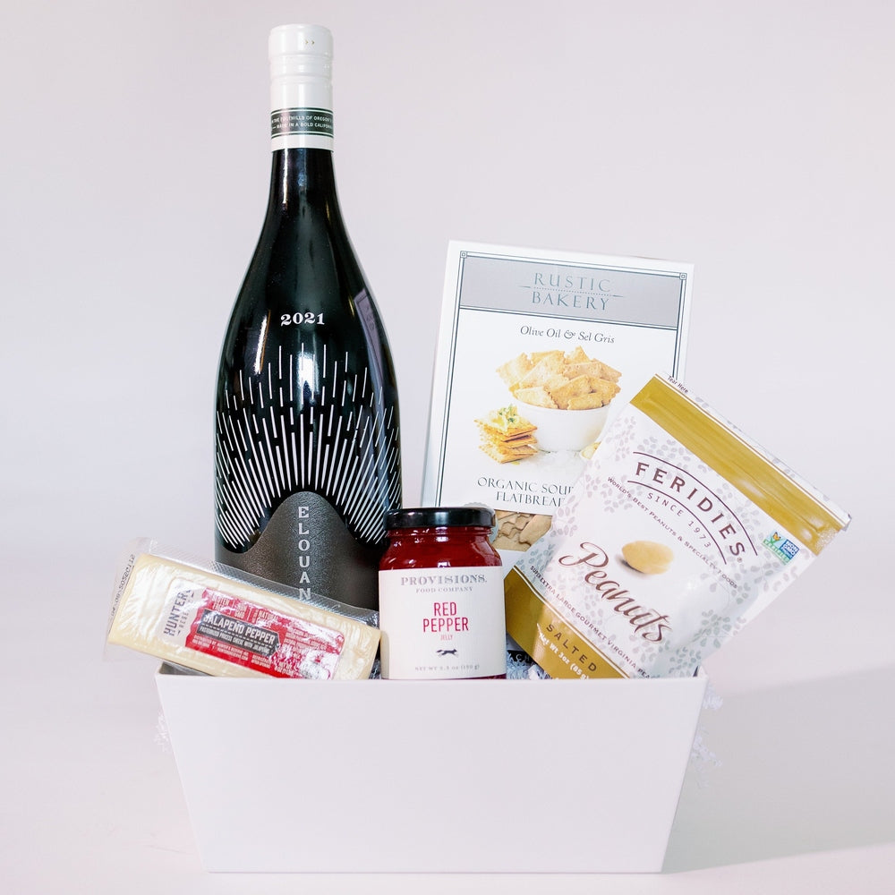 Studio photograph of a gourmet gift basket in a white container featuring a 2021 Elouan wine bottle with a white radiating pattern, a bright red jar of Provisions pepper jelly, and a wedge of Hunter Jalapeno Pepper cheese