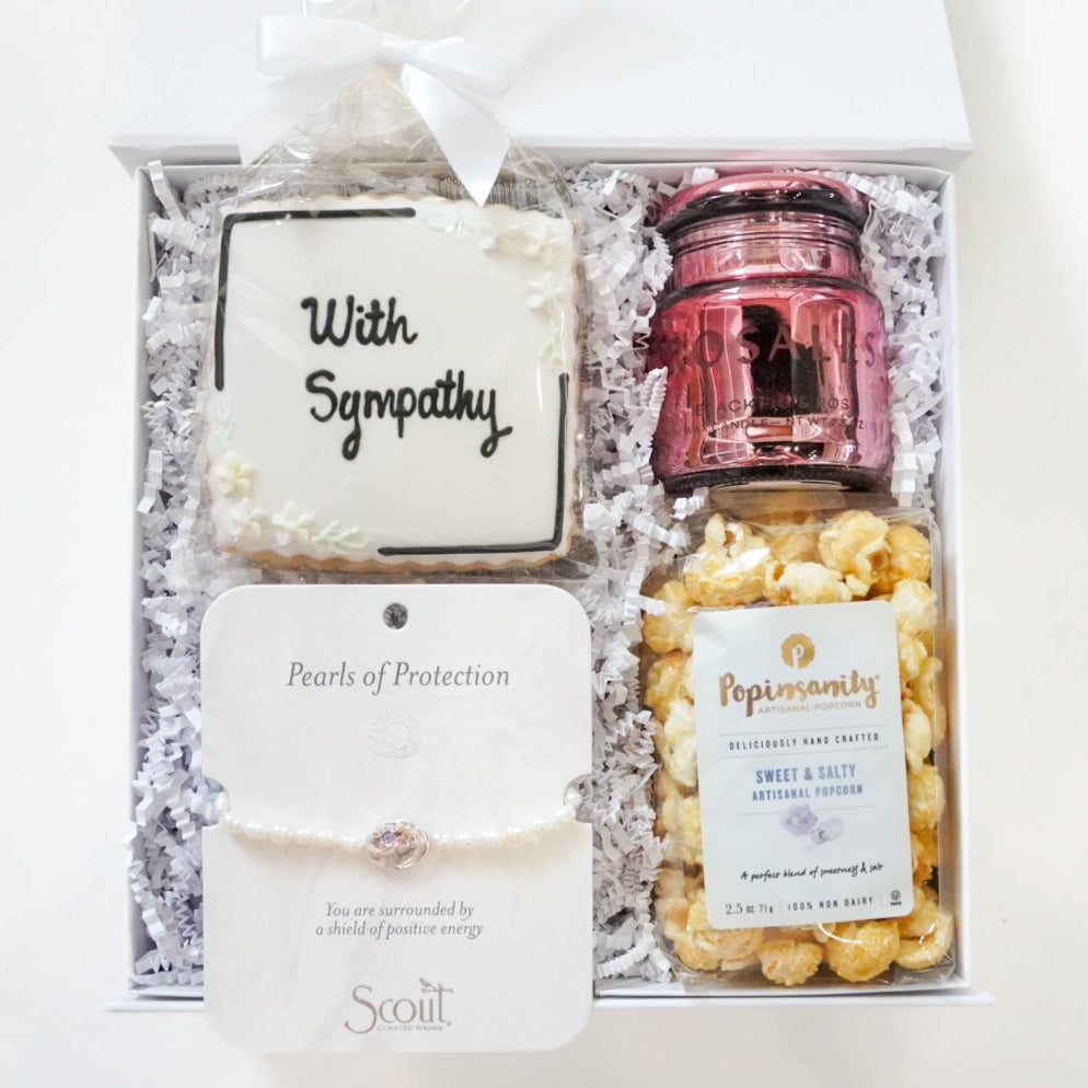 Photograph of a curated sympathy gift box featuring a white iced cookie with With Sympathy script, a Scout Curated Wears pearl bracelet, a bag of Popinsanity gourmet popcorn, and a metallic pink candle jar nestled in white shredded paper filler