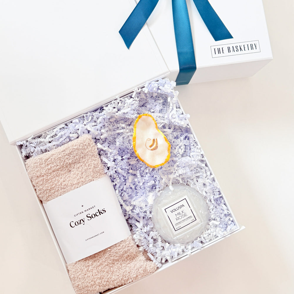Gift set with cozy socks, candle, and bottle labeled 'The Merchant' on a light background