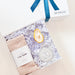 Gift set with cozy socks, candle, and bottle labeled 'The Merchant' on a light background
