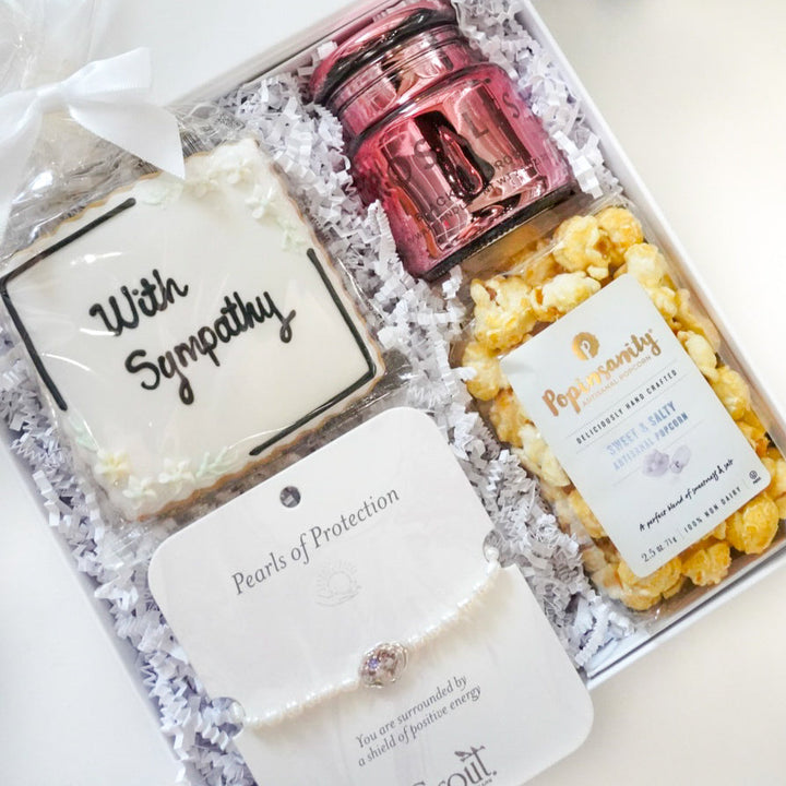 Photograph of a curated sympathy gift box containing a white frosted sugar cookie with black calligraphy, a metallic pink candle jar, Popinsanity popcorn, and a Scout Curated Wears pearl bracelet on white crinkle paper