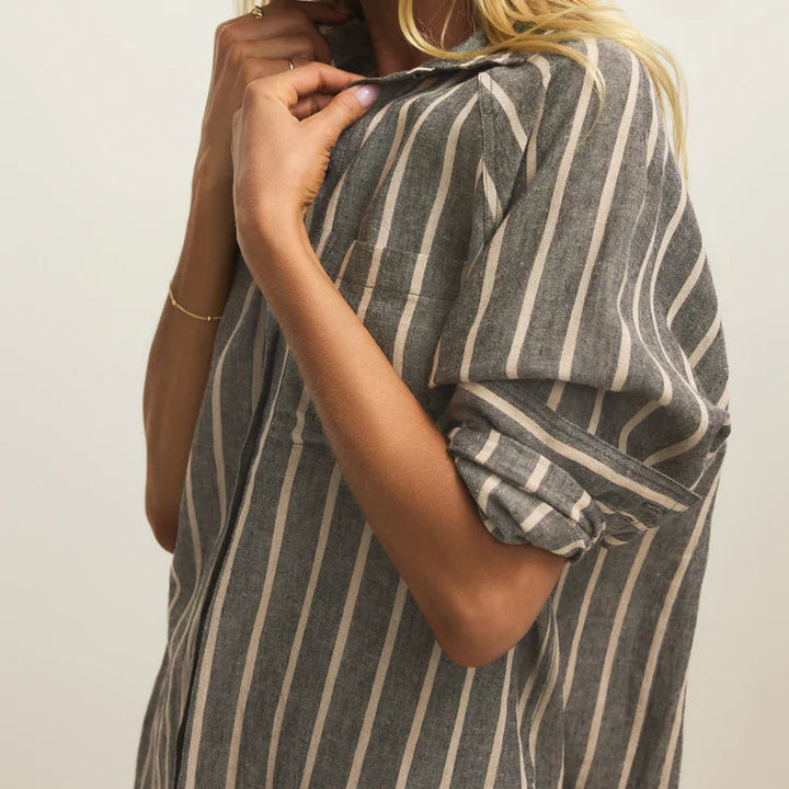Photograph of a woman wearing a gray and white vertical striped shirt in a studio featuring rolled-up sleeves and delicate gold jewelry