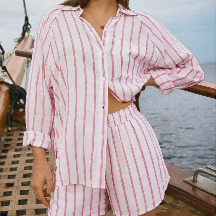 Photograph of a woman wearing a pink and white vertically striped linen shirt and shorts set on a wooden sailboat featuring a front chest pocket and elastic waistband overlooking the open ocean