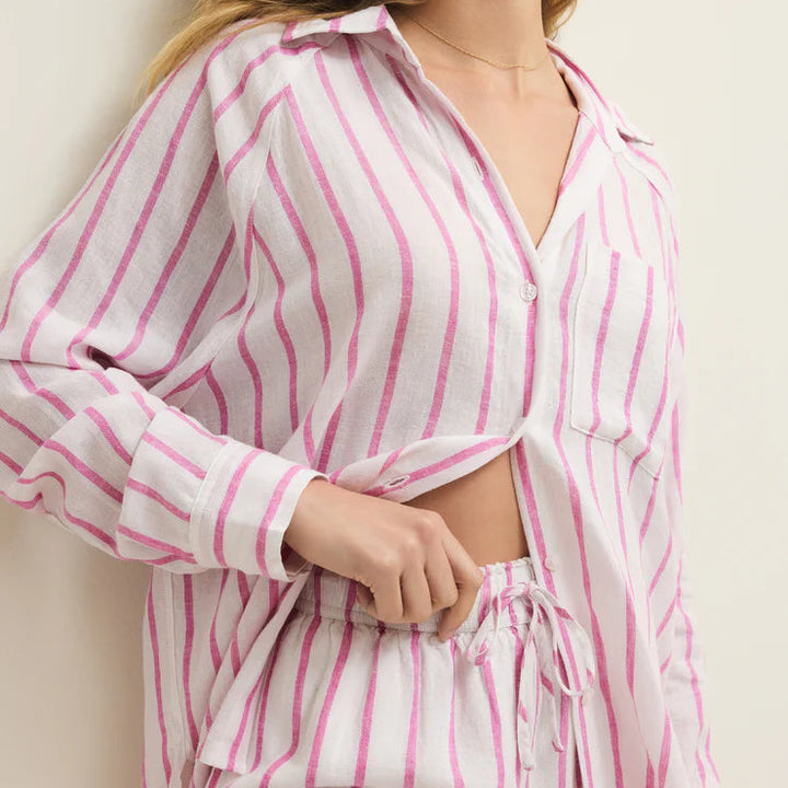 Digital photograph of a woman wearing a pink and white striped linen lounge set in an indoor setting, featuring a button-down shirt with a chest pocket and matching drawstring shorts accented by a delicate gold necklace