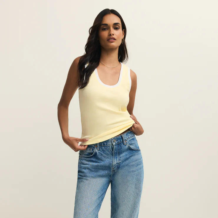 Digital photograph of a woman modeling in a studio wearing a pale yellow ribbed ringer tank top with white trim and faded blue high-waisted denim jeans