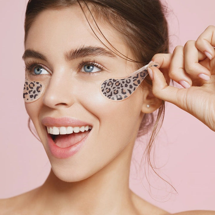 Digital studio photograph of a smiling woman with blue eyes against a pink background wearing leopard print under-eye patches and a small pearl earring