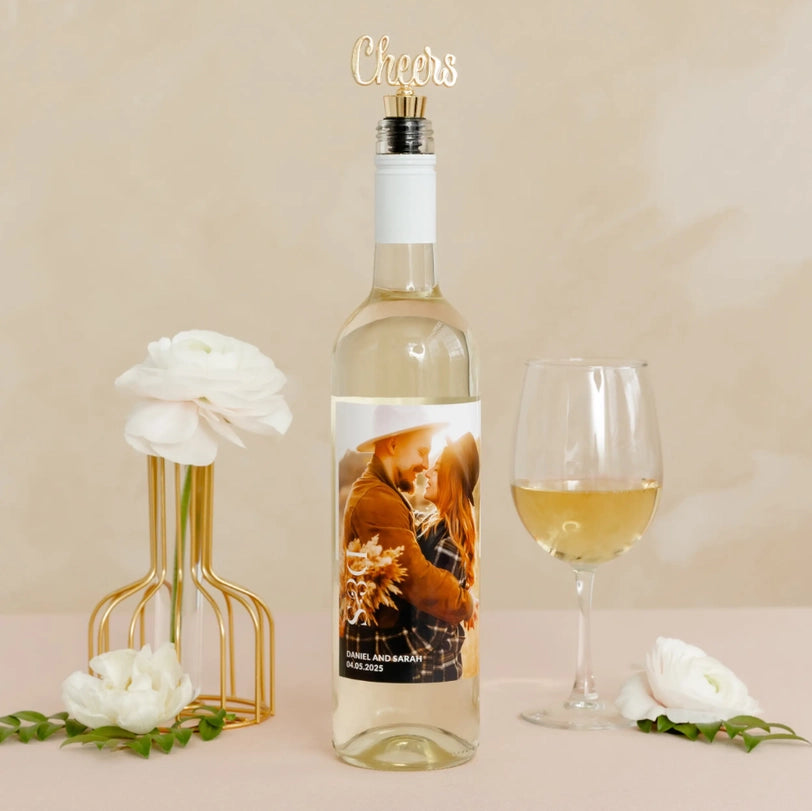 Photograph of a personalized white wine bottle featuring a custom photo label for Daniel and Sarah, a gold cursive Cheers topper, and a glass of wine next to white ranunculus flowers in a gold wire vase