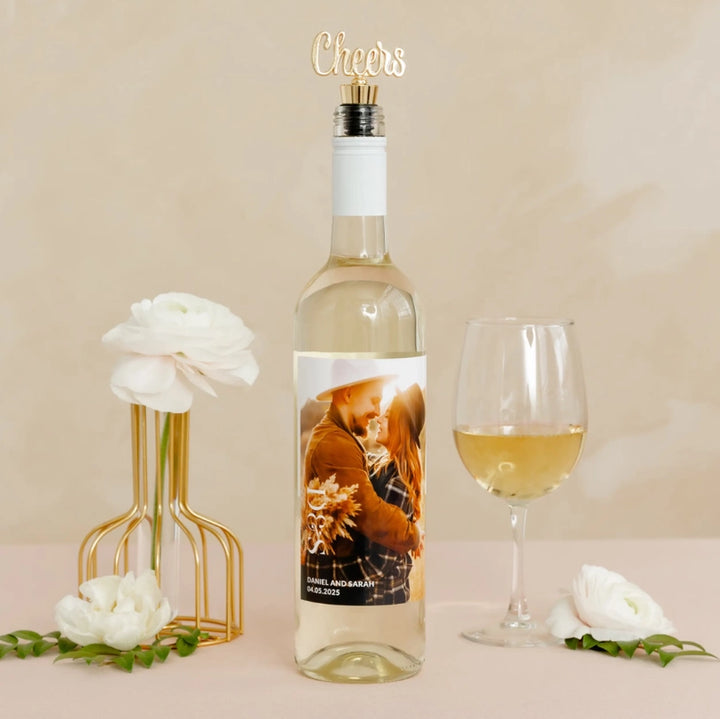 Photograph of a personalized white wine bottle featuring a custom photo label for Daniel and Sarah, a gold cursive Cheers topper, and a glass of wine next to white ranunculus flowers in a gold wire vase