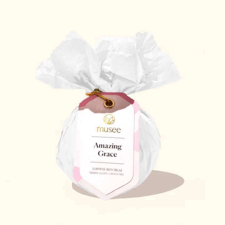 Product photograph by Musee of an Amazing Grace surprise bath balm wrapped in white crinkled tissue paper featuring a pink-accented tag with gold lettering and a gold metallic cord on a cream background