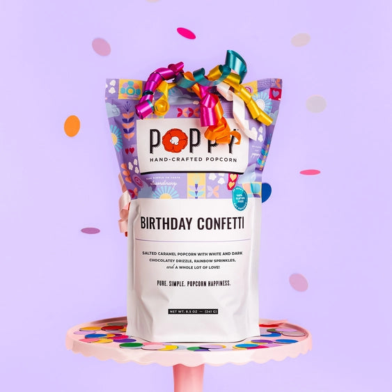 Studio photograph by Poppy Hand-Crafted Popcorn of a Birthday Confetti bag on a pink scalloped cake stand against a lavender background with vibrant curling ribbons and falling multi-colored confetti