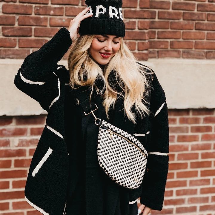 Digital photograph of a blonde woman posing in front of a red brick wall wearing a black and white fuzzy sherpa jacket, a checkered crossbody bag, and a black knit beanie with white lettering