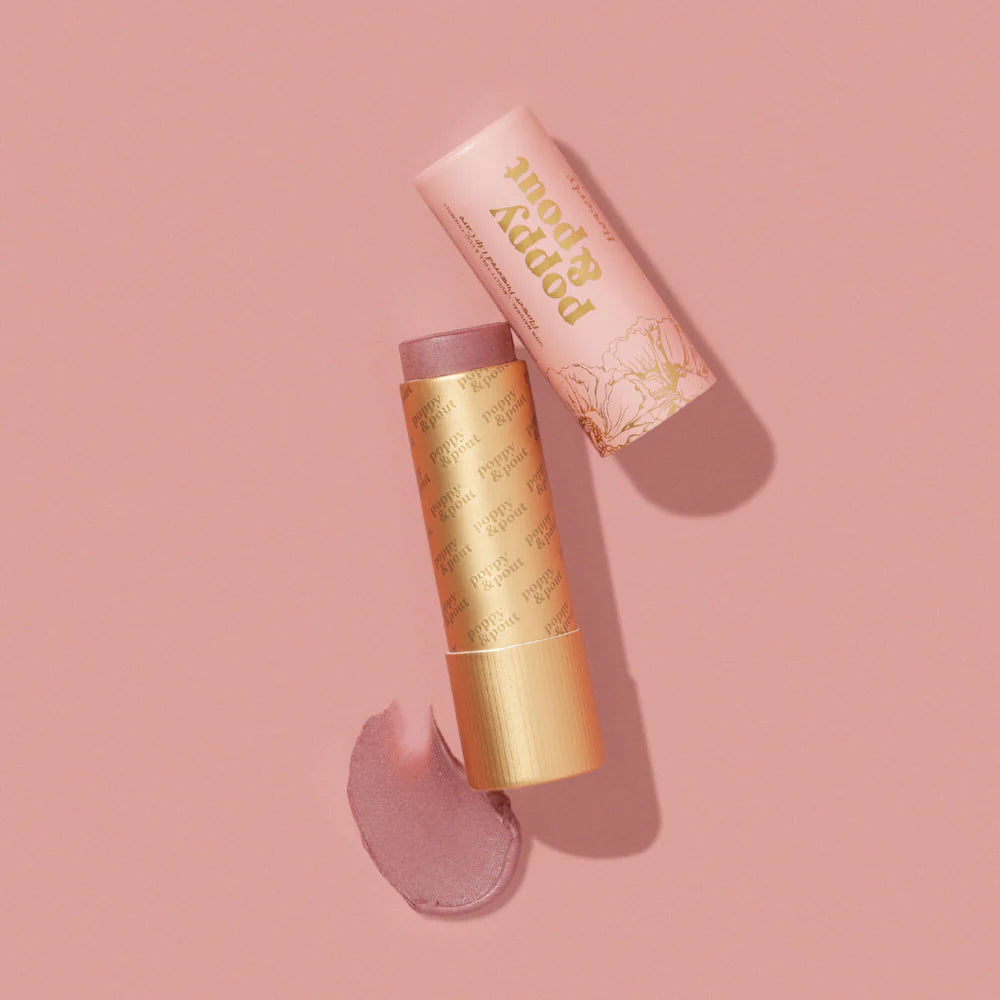 Product photography of Poppy &amp; Pout tinted lip balm on a monochromatic pink surface featuring an open cardboard tube with gold floral illustrations, a shimmery mauve balm stick, and a textured swatch of the product