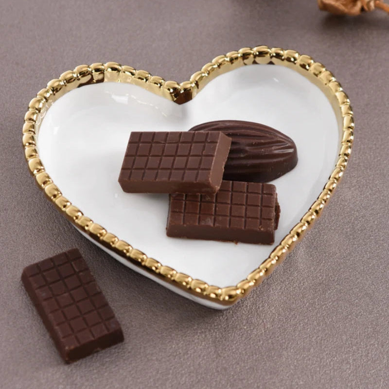 Still life photograph of a white heart-shaped ceramic dish with a shimmering gold beaded rim holding grid-patterned chocolates and a dark cocoa oval on a textured earth-toned background with one chocolate bar placed intentionally to the side