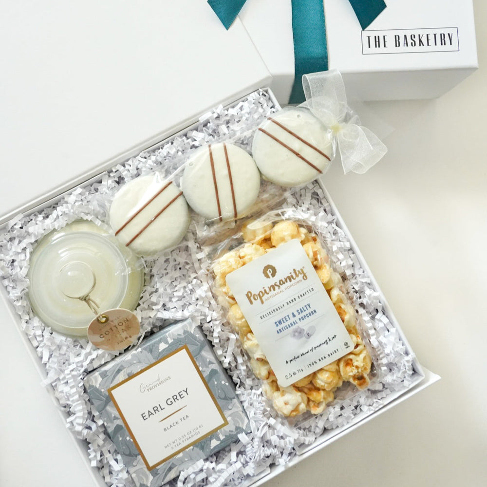 Photograph by The Basketry of a curated gourmet gift box featuring Popinsanity artisanal popcorn, Grand Provisions Earl Grey tea, and three white chocolate-dipped cookies with chocolate stripes nestled in white crinkle paper with a dark teal ribbon