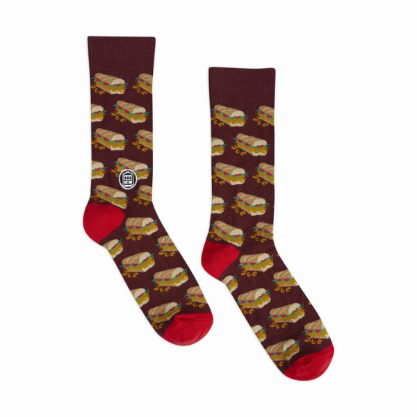 Woven crew socks featuring a repeating pattern of New Orleans dressed po-boy sandwiches against a maroon background with a white St. Charles streetcar logo and vibrant red heel and toe accents