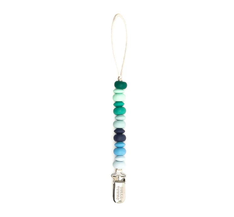 Silicone and metal pacifier clip by Bella Tunno featuring a vertical string of teal, navy, and mint green beads with a silver metal clip engraved with the brand name on a white background