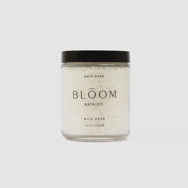 Studio photograph of Bloom Bath Co. Wild Rose bath soak in a clear glass jar against a neutral grey background featuring a matte black screw-top lid, coarse white crystalline salts, and minimalist black serif typography