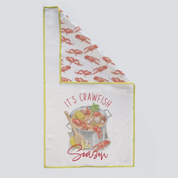 Illustrated waffle-weave kitchen towel showcasing a central crawfish boil pot with corn and mushrooms, red script reading IT'S CRAWFISH Season, and a vibrant yellow border with a folded-over corner revealing a red crawfish pattern