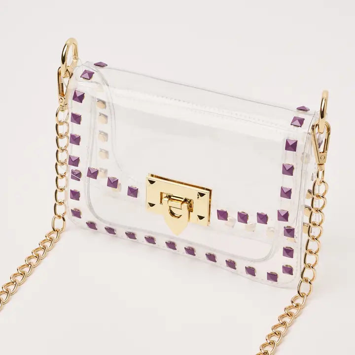Product photograph of a transparent PVC crossbody bag against a plain white studio backdrop featuring purple pyramid studs along the border and polished gold-tone hardware including a turn-lock clasp and chain strap