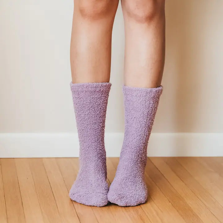 Photograph of a person's feet and lower legs wearing fuzzy lavender-colored socks with a plush chenille texture while standing on a light-toned hardwood floor against a neutral wall with a white baseboard