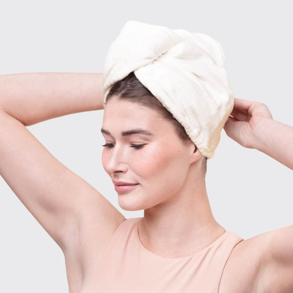 Studio photograph of a woman in a minimalist studio wearing a textured white microfiber hair towel wrap and a soft peach-colored tank top against a neutral gray backdrop