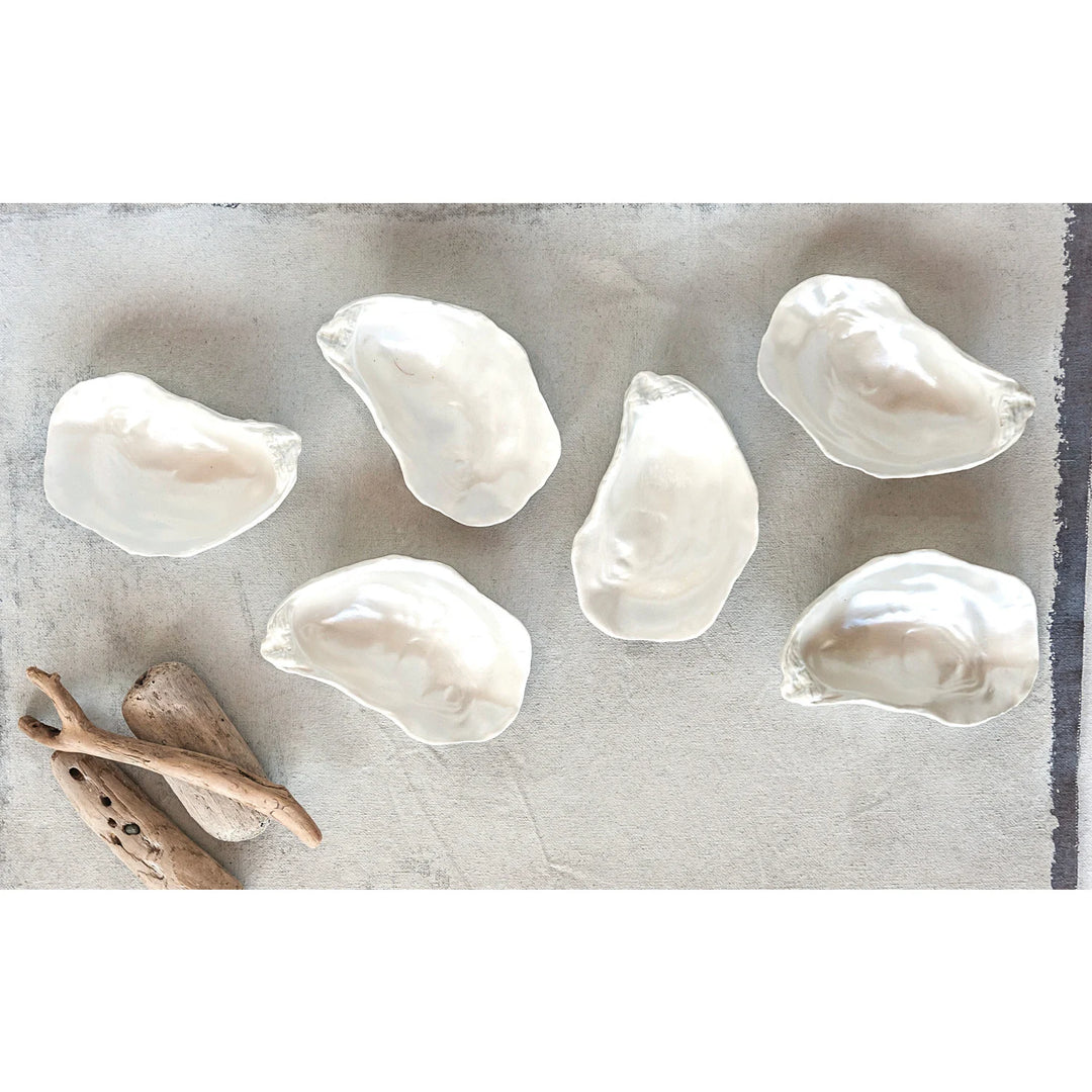 Ceramic art featuring six iridescent oyster shell bowls arranged on a textured linen background accompanied by weathered driftwood with organic ridged edges and a luminous pearlescent glaze