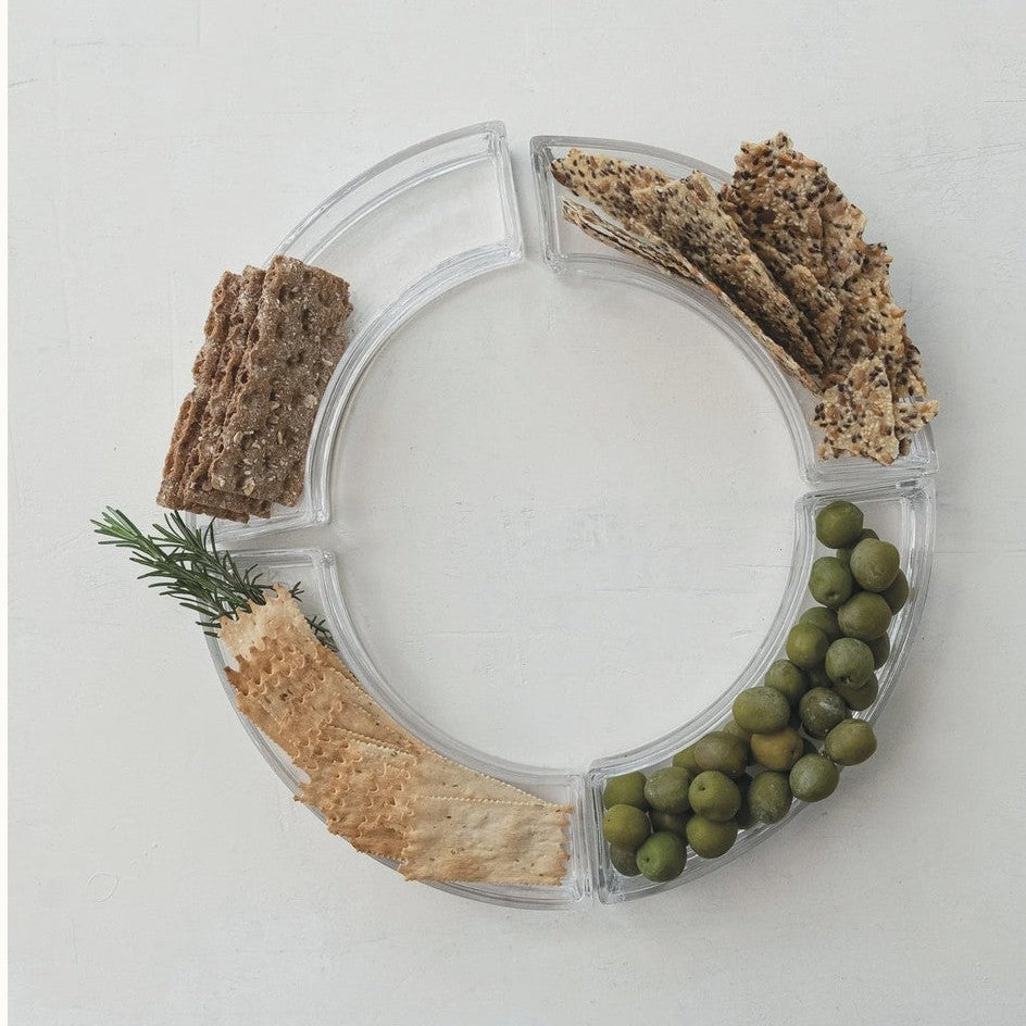 Still life photograph of a clear circular segmented tray on a white textured background featuring green olives, multi-seed crackers, and a fresh sprig of rosemary
