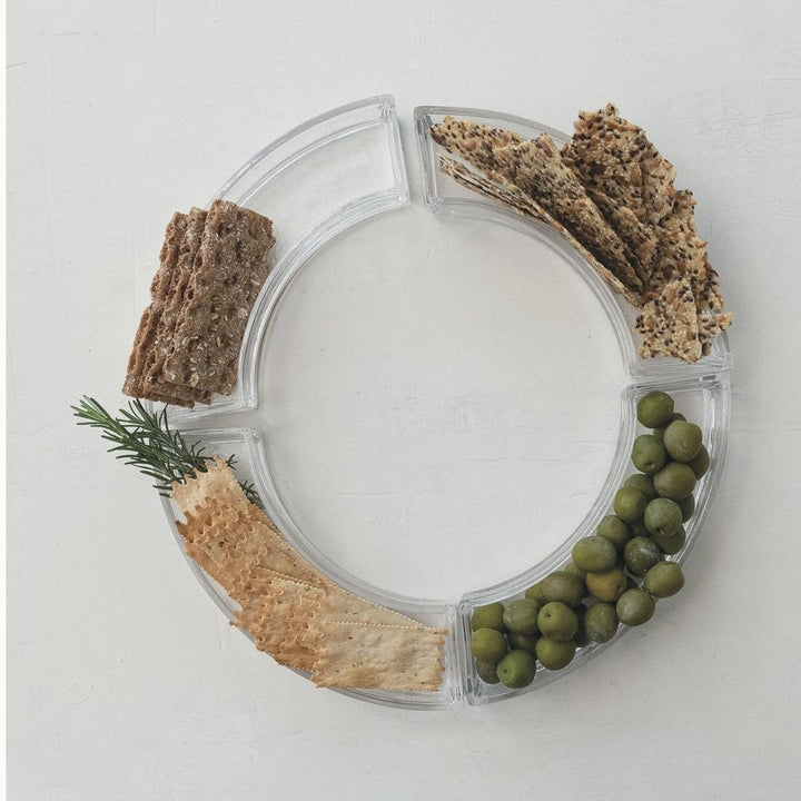 Still life photograph of a clear circular segmented tray on a white textured background featuring green olives, multi-seed crackers, and a fresh sprig of rosemary