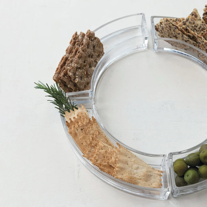 Photograph of a modular clear acrylic circular serving tray on a white textured surface featuring green olives, various artisanal crackers, and a fresh sprig of green rosemary