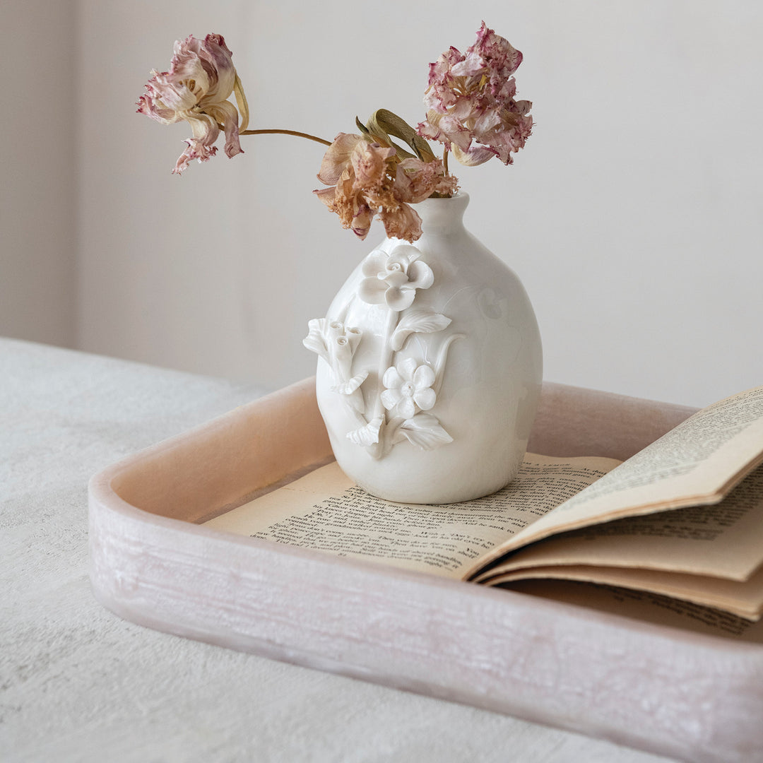 Still life photograph of a white ceramic vase with intricate 3D floral relief and withered pink dried flowers positioned on an open vintage book inside a textured blush pink tray on a neutral indoor surface