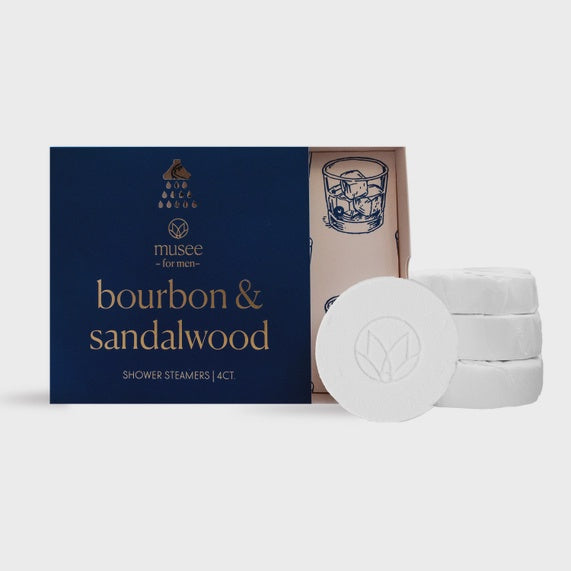 Product photograph by Musée featuring a navy blue box of bourbon and sandalwood shower steamers for men accompanied by white circular tablets with an embossed lotus logo and a line-art whiskey glass illustration on a minimalist background