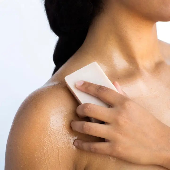 Digital photograph of a person with deep-toned skin applying a cream-colored bar of soap to their wet shoulder against a minimalist white background, highlighting glistening water droplets and moisturized skin texture