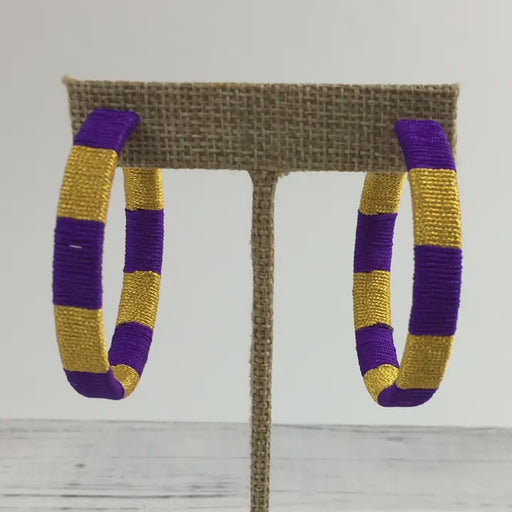 Handcrafted thread-wrapped hoop earrings featuring vibrant alternating purple and gold stripes displayed on a textured burlap jewelry stand against a plain white background