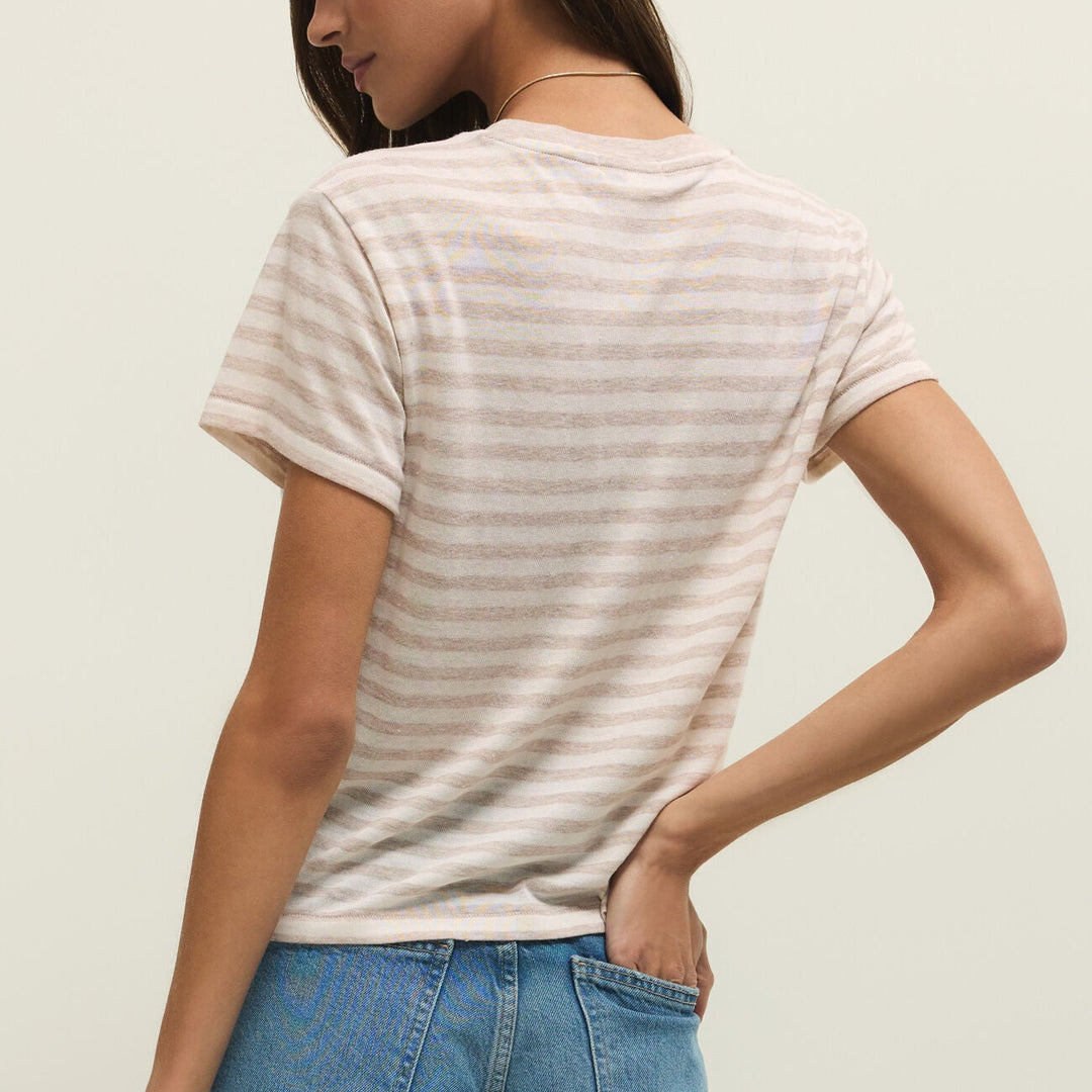 Studio photograph of a woman from a rear-facing view wearing a beige and white horizontal striped t-shirt and blue denim jeans with her hand in a pocket and a thin gold necklace against a plain studio background