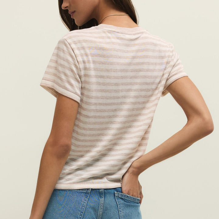 Studio photograph of a woman from a rear-facing view wearing a beige and white horizontal striped t-shirt and blue denim jeans with her hand in a pocket and a thin gold necklace against a plain studio background