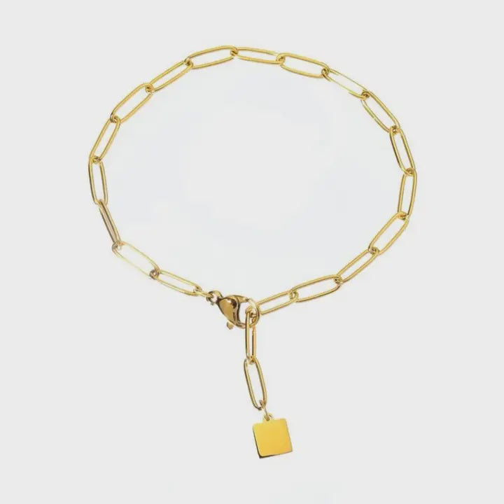 Gold jewelry paperclip chain bracelet on a light gray background featuring polished elongated oval links, a lobster claw clasp, and a small square gold charm dangling from the extender chain