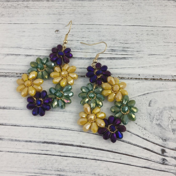Handcrafted beaded jewelry featuring a pair of floral drop earrings with shimmering purple, golden-yellow, and sage green faceted beads resting on a rustic white-washed wooden surface