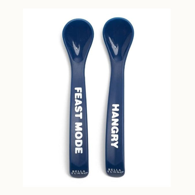 Silicone baby spoons by Bella Tunno featuring two navy blue utensils on a white background with prominent white block lettering reading FEAST MODE and HANGRY above the brand logo at the rounded base