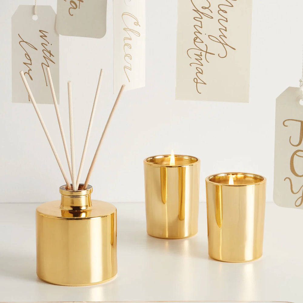 Professional product photograph of a gold home fragrance set on a white tabletop featuring a reflective metallic reed diffuser and two lit votive candles positioned before hanging holiday tags with gold calligraphy